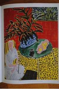 This picture is called Interior with Black Fern and is an oil painting. It is a very colourful painting where abstraction and realisms are combine together. A white dressed woman in the bottom left corner is sitting on a yellow chair in a living room. In the centre is a green table with a black cup, a pink plate with oranges inside and a big blue pot with white dots on it. Inside this pot is a large dark green fern that occupy a quarter of the painting. The ground is yellow with black dots and the wall in front is chequered red. At the upper right corner is a window and behind this window there are several plants visible.