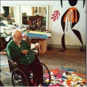 This is a photo of Henri Matisse himself during the last years of his life in his house in Vence in France that was as well his atelier. Matisse is seating in a wheelchair with two large wheels in the front and two small in the back, only the large wheels are visible. He is wearing a black pants and a green pullover. He have white hair and a white beard and he is holding somthing  with his two hands. There are a lot of coloured paper cutting on the floor and they is one big cut out fixed on the wall representing a black women with orange skirt and a pink leaves on white background. Behind him there is a lot of working and painting materials.